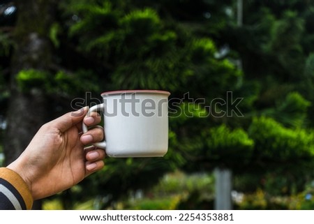 A hand holding an enamel mug with out of focus tree on the background at a park, enamel mug mockup image