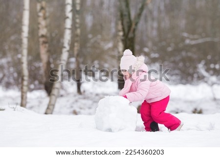Little girl in pink warm clothes rolling white big snowball at nature park. Cute 3 years old toddler enjoying cold winter day. Side view.