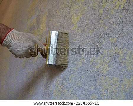 applying a layer of adhesive or primer to a textured wall with a wide, hard bristled brush close-up, a gloved hand of a painter holding a brush and applying mortar to the surface of the wall Royalty-Free Stock Photo #2254365911
