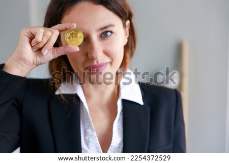 Smiling young woman covering the eye with bitcoin coin currency in the office. The focus is on the coin