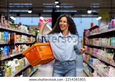 Portrait of a happy woman shopper in a supermarket, a Hispanic woman with a basket of goods smiles with pleasure and dances among the shelves with goods, a satisfied shop customer. Royalty-Free Stock Photo #2254381801