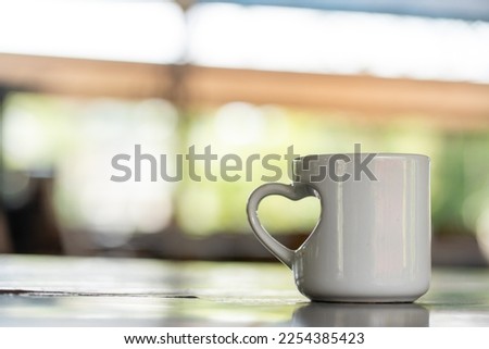 A coffee mug with a love shaped handle standing on the top of a table with blurred out bright background, coffee mug mockup image
