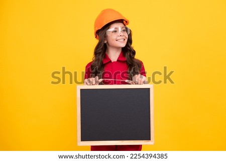 Child girl builder in hard hat helmet. Teenage girl worker hold blackboard isolated on yellow background. Kids renovation concept. Copy space, mock up.
