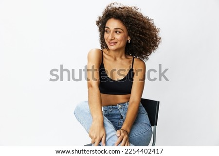 Woman with curly afro hair model poses on a chair against a white background, free movement and dance, looking into the camera, copy space