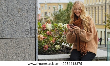 Caucasian woman 20s girl in glasses sitting outdoors in city browsing scrolling smartphone smiling female lady shopping online with phone mobile store app chatting with friend in cell social media