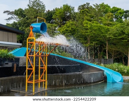 A blue water slide next to a water pouring large blue bucket on top of a colorful metal scaffolding at the side of a children pool. Royalty-Free Stock Photo #2254422209