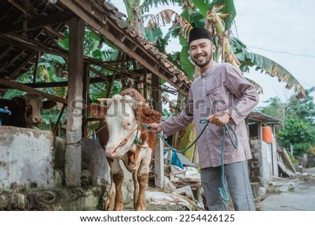 a moslem man with skullcap standing in front of the cow's stable and holding the cow's bridle that standing next to him Royalty-Free Stock Photo #2254426125