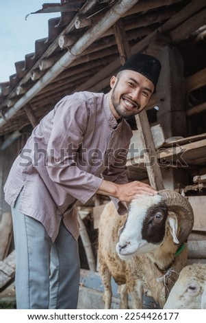 a moslem man with skullcap standing in front of the goat's stable and stroking the goat's head that standing next to him Royalty-Free Stock Photo #2254426177