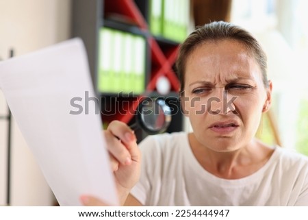 Incredulous middle age woman looking on document trough magnifying glass close-up. Concept of eye diseases and people incredulity.