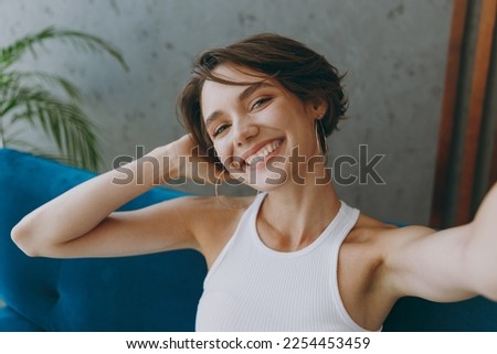Young smiling woman wear white tank shirt do selfie shot pov on mobile cell phone sit on blue sofa stay at home hotel flat rest relax spend free spare time in living room indoor People lounge concept