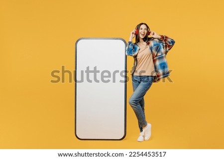 Full body young woman wear blue shirt beige t-shirt headphones listen music big huge blank screen mobile cell phone smartphone with workspace copy space mockup area isolated on plain yellow background