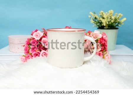 A white blank enamel mug standing out on top of a fluffy white mat with some flower behind it, enamel mug mockup image