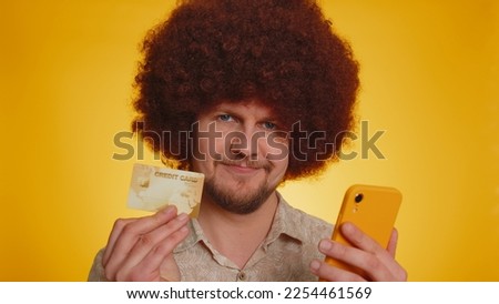 Sincere man with lush afro hairstyle customer using credit bank card and smartphone while transferring money purchases online shopping, payment. Finance and internet. Young guy on yellow background