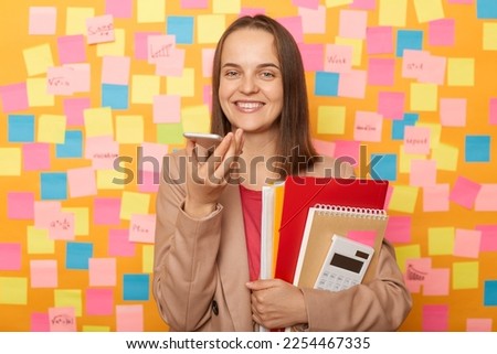 Image of charming cute beautiful woman wearing beige jacket, holding her documents, recording voice message or commands standing against yellow wall with colorful stickers.