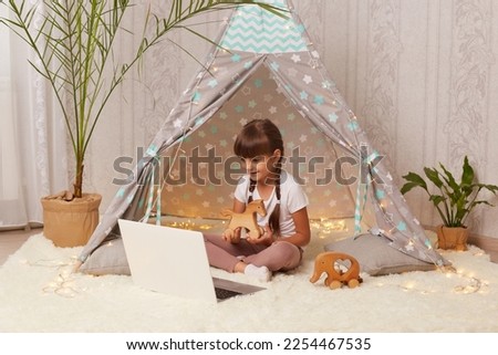 Indoor shot of satisfied delighted little girl with pigtails lying in peetee tent with laptop and having online conversation with her friend, showing her new wooden horse.