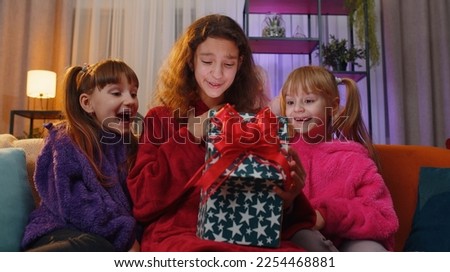 Happy teen girl and toddler child sisters opening Christmas gift box with excited surprised face. Female three children kids siblings or friends holding light glowing birthday present at home playroom