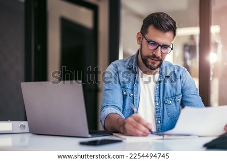 Satisfied young man with glasses sitting at a desk and doing paperwork at his workplace. Royalty-Free Stock Photo #2254494745