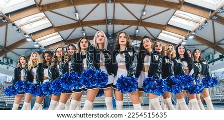 Front view of a group of cheerleaders in black and white uniforms with blue shiny pom-poms in their hands posing in sports hall. High quality photo