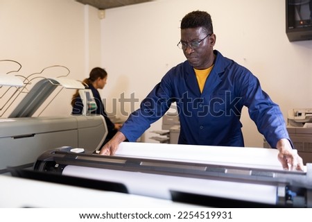 Serious middle-aged African American man in uniform loading large format paper in a plotter in the print shop