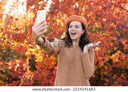 Attractive young smiling woman walking in autumn park having video call or taking selfie pictures using smartphone, wearing styling beret, happy mood. Copy space