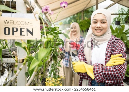 Beautiful young muslim wearing hijab is standing in greenhouse with we are open sign smiling welcoming buyers and using pruning shears. Business and floristry concept.