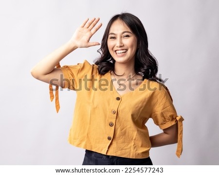 A studio portrait of a young beautiful Indonesian Asian woman in a casual suit waving her hand as if saying hai. Isolated on white background. Royalty-Free Stock Photo #2254577243