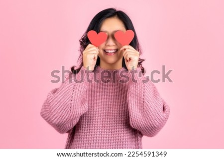 Cute asian woman wearing pink sweater holding red love hearts over eyes standing isolated on pink background, love and caring people concept