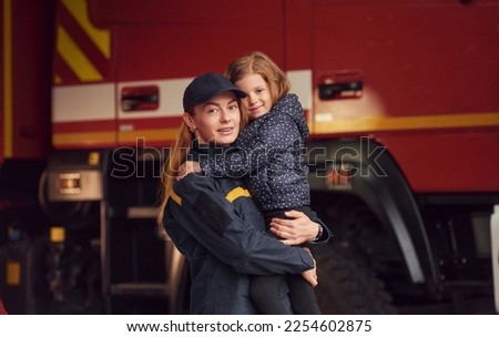 Firefighter woman in uniform is holding a little girl in the hands.