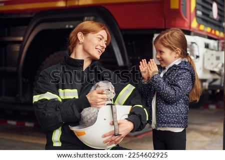Cat is sitting in the helmet, kid is happy, clapping her hands. Firefighter woman in uniform is with a little girl.