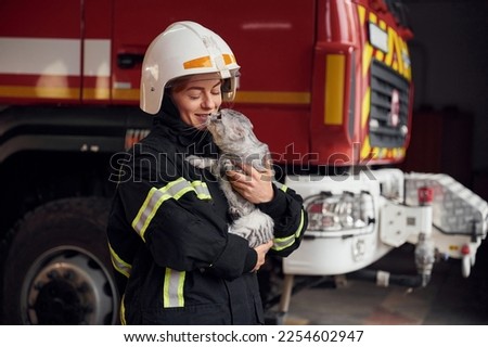 Standing against big truck and holding beautiful cat. Woman firefighter in uniform is at work in department.