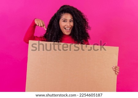 Curly-haired woman pointing at a sign on a pink background, studio shot