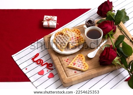 Romantic breakfast, wooden tray with freshly baked homemade heart shaped Belgian waffles, cup of tea or coffee and red rose on red white table background. Valentine's Day breakfast surprise idea.