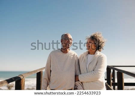 Mature couple smiling happily while taking a walk along a wooden foot bridge at the beach. Romantic elderly couple enjoying a refreshing seaside holiday after retirement. Royalty-Free Stock Photo #2254633923