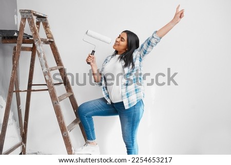 Wide shot of young Indian woman singing with paint roller while painting wall