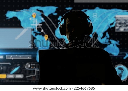 Silhouette of a military man in headphones at a laptop against the background of a world map, contour lighting. Concept: collection of confidential information, surveillance and control of people. Royalty-Free Stock Photo #2254669685