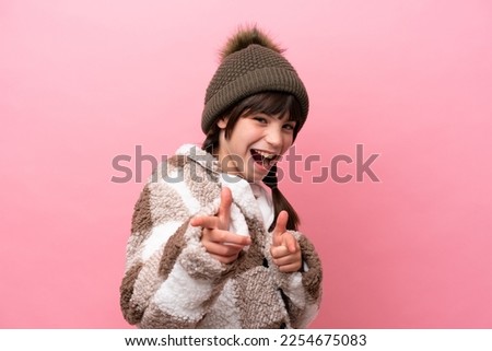 Little caucasian girl with winter jacket isolated on pink background pointing to the front and smiling