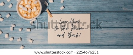 LOVE YOU TO THE MOON AND BACK text on valentine card inscription positive quote phrase. Greeting card with white cup of coffee and marshmallows on wooden blue background. holiday concept. 