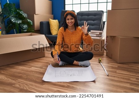 Young hispanic woman sitting on the floor at new home looking at blueprints doing ok sign with fingers, smiling friendly gesturing excellent symbol 