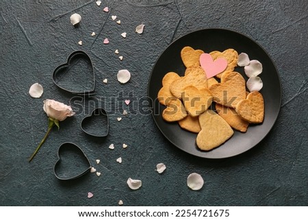 Composition with plate of tasty heart shaped cookies, cutters and rose petals on dark color background. Valentines Day celebration