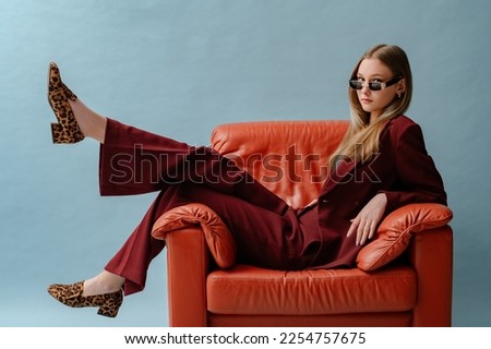 Fashionable confident woman wearing elegant marsala color suit, sunglasses, leopard print loafer shoes, posing, sitting in brown leather armchair. Studio fashion portrait. Copy. empty space for text Royalty-Free Stock Photo #2254757675