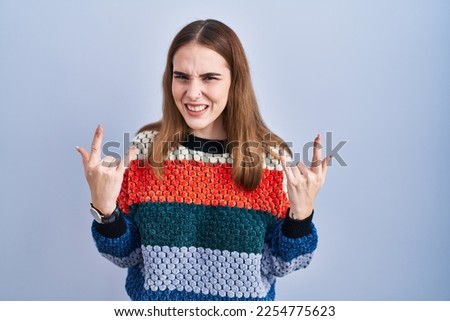 Young hispanic girl standing over blue background shouting with crazy expression doing rock symbol with hands up. music star. heavy music concept. 