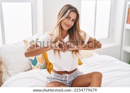 Young blonde woman doing heart gesture sitting on bed at bedroom