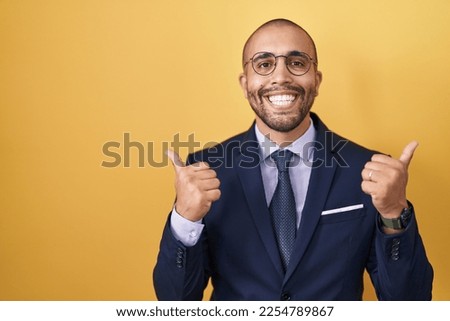 Hispanic man with beard wearing suit and tie success sign doing positive gesture with hand, thumbs up smiling and happy. cheerful expression and winner gesture. 