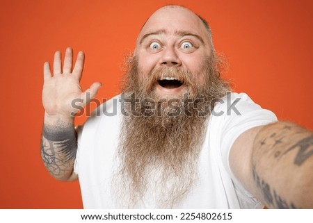 Close up fat pudge obese chubby overweight tattooed blue-eyed bearded man in white t-shirt do selfie shot on mobile phone waving hand greet hello gesture isolated on orange background studio portrait