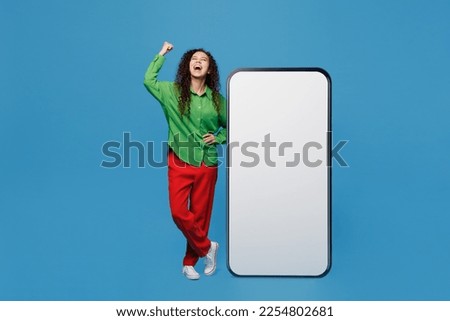 Full body young woman of African American ethnicity wear green shirt near big huge blank screen mobile cell phone with workspace copy space mockup do winner gesture isolated on plain blue background
