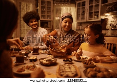 Happy Muslim grandmother serving food to her granddaughter during family dinner at dining table. Royalty-Free Stock Photo #2254805311