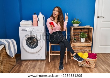 Young hispanic woman sitting waiting for laundry using smartphone smiling with happy face winking at the camera doing victory sign with fingers. number two. 