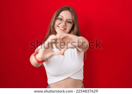 Young caucasian woman standing over red background smiling in love doing heart symbol shape with hands. romantic concept. 