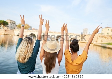 Back view of happy young women friends sitting with raised hands on the lake in the resort town. Concept of tourism and friendship. High quality photo