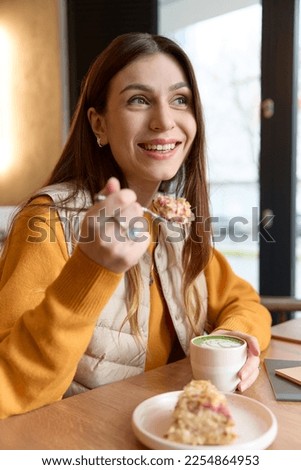 Portrait of a smiling woman eating a piece of cake dessert and drinking green matcha, sitting at a table in a cafe. Beautiful caucasian brunette woman sitting in a light cafe indoors. Royalty-Free Stock Photo #2254864953